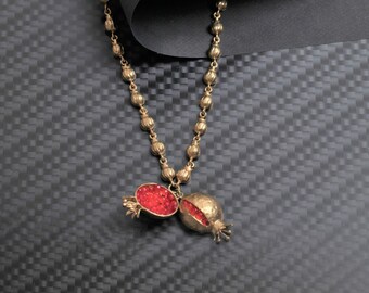 Peut inclure: Un collier en chaîne dorée avec un pendentif en forme de grenade. Le pendentif est ouvert et montre des graines de grenade rouges à l'intérieur.