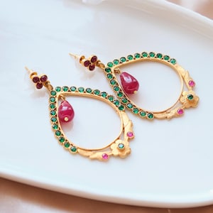 May include: Gold-toned teardrop earrings with green and red gemstones. The earrings feature a floral design and a dangling red gemstone. The earrings are displayed on a white surface.