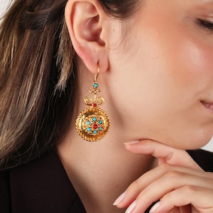 May include: Gold earrings with a round turquoise and red stone design. The earrings have a dangling design with a small loop at the top.