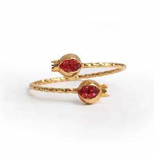 May include: A gold-colored ring with a hammered texture, featuring two pomegranate-shaped ends. Each pomegranate holds a cluster of small, red gemstones. The ring's design wraps around the finger, creating a unique and elegant look.