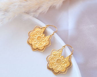 May include: A pair of gold-colored filigree earrings. Each earring features a floral design with intricate cut-out details. The earrings have a hook closure and are displayed on a white surface.