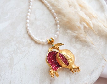 Peut inclure: Un collier de perles avec un pendentif en or en forme de grenade. Le pendentif représente deux moitiés de grenade, avec un intérieur rouge semblable à une gemme et un extérieur doré. Le collier est posé sur une surface en marbre blanc.