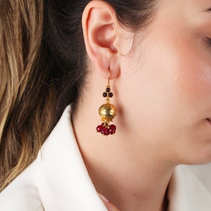 Peut inclure: Boucles d'oreilles pendantes dorées avec un centre sphérique texturé et des grappes de perles rouges. Les boucles d'oreilles ont une fermeture à crochet et de petits bijoux rouges en haut. Le design est élégant et accrocheur.