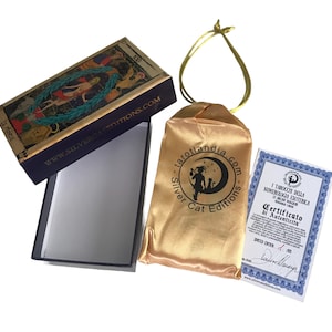 May include: A tarot deck box with a gold and blue design. The box is open and the lid is flipped over. The lid features a tarot card with a woman standing in a forest. The box is sitting on a white surface. The box is next to a gold satin bag with a black logo that says "tarotlandia.com - Silver Cat Editions". The bag is tied with a gold ribbon. A certificate of authenticity is also visible. The certificate is printed on white paper with a blue border and features a black and white illustration of a tarot card. The text on the certificate reads "I Tarocchi della Numerologia Esoterica 22 Arcani Maggiori - Certificato di Autenticita' - Limited Edition - No. 200".