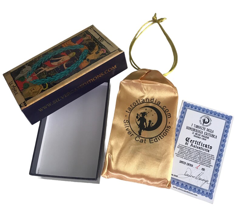 May include: A tarot deck box with a gold and blue design. The box is open and the lid is flipped over. The lid features a tarot card with a woman standing in a forest. The box is sitting on a white surface. The box is next to a gold satin bag with a black logo that says "tarotlandia.com - Silver Cat Editions". The bag is tied with a gold ribbon. A certificate of authenticity is also visible. The certificate is printed on white paper with a blue border and features a black and white illustration of a tarot card. The text on the certificate reads "I Tarocchi della Numerologia Esoterica 22 Arcani Maggiori - Certificato di Autenticita' - Limited Edition - No. 200".