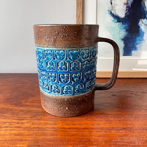 May include: A ceramic mug with a brown top and base, and a vibrant blue floral pattern in the middle. The mug has a large, curved handle and a textured surface. The mug is sitting on a wooden surface.