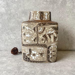 May include: A rectangular ceramic vase featuring a brown and white glaze with intricate, segmented panels depicting stylized fish, floral motifs, and geometric patterns. The surface has a textured, hand-drawn appearance with dark brown outlines.