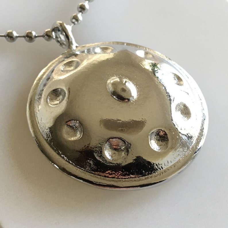 May include: A silver handpan pendant necklace. The pendant is round and has eight indentations. The necklace is made of a silver chain.