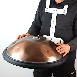 Peut inclure: Un instrument de musique handpan de couleur bronze avec un bord en caoutchouc noir. L'instrument est maintenu par un harnais blanc. Le handpan a plusieurs fossettes sur sa surface. Le fond est blanc uni.