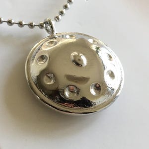 May include: A silver handpan pendant necklace. The pendant is round and has a hammered finish. It has a small, raised center dome and eight smaller, evenly spaced domes around the edge.