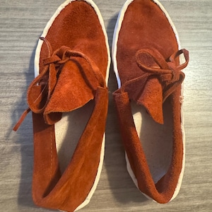 May include: A pair of rust-colored suede moccasins with white soles and laces. The shoes have a soft, textured appearance and a classic design. The laces are tied in a bow at the top of each shoe.