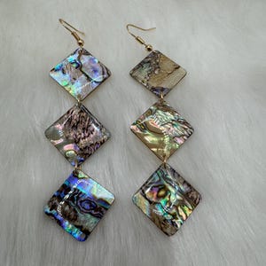 May include: A pair of long, dangling earrings with three iridescent, square-shaped pieces of abalone shell. The earrings are gold-toned and have a simple hook closure.