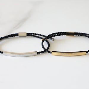 Puede incluir: Dos pulseras de cordón negro con barras de metal plateado y dorado. La barra plateada tiene una inscripción y la barra dorada tiene la inscripción "DELTA ZETA".
