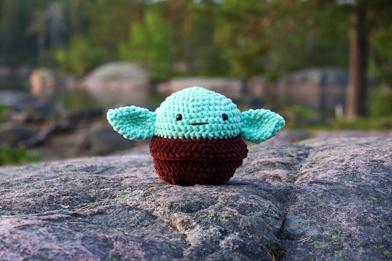 May include: A crocheted plush toy resembling a popular fictional character. The toy is green with brown accents and has a friendly expression.