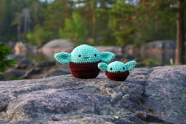 Pu&ograve; includere: Due giocattoli di peluche all'uncinetto a forma di un popolare personaggio di fantasia. Il giocattolo pi&ugrave; grande &egrave; verde con accenti marroni e il giocattolo pi&ugrave; piccolo &egrave; verde con accenti marroni. I giocattoli sono seduti su una roccia grigia.