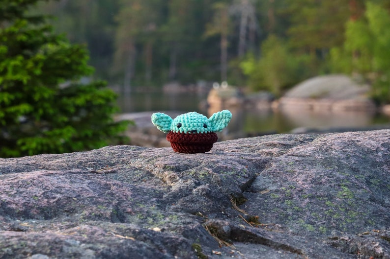 Pu&ograve; includere: Un piccolo giocattolo di peluche verde e marrone all'uncinetto che ricorda un personaggio della saga di Star Wars, seduto su una roccia grigia con uno sfondo sfocato di alberi e un lago.