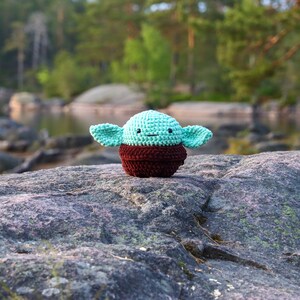 May include: A small crocheted plush toy of a green alien character with large ears, brown eyes, and a brown vest. The toy is sitting on a grey rock with a blurred background of trees and water.