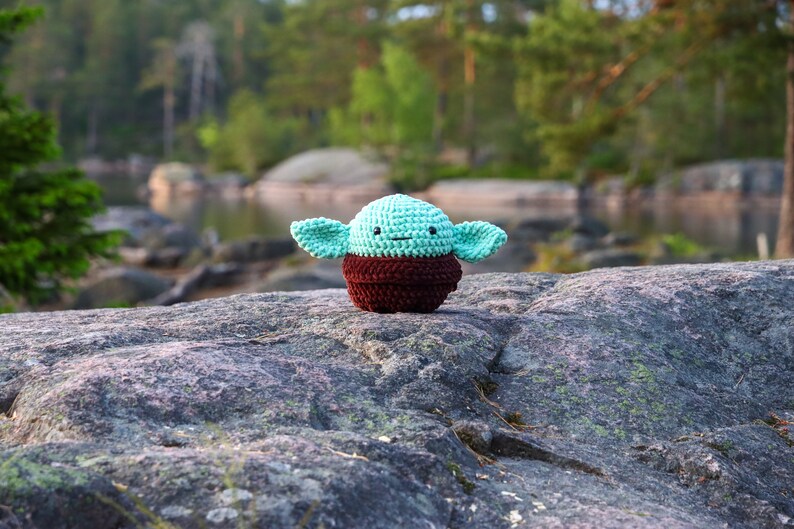 May include: A small crocheted plush toy of a green alien character with large ears, brown eyes, and a brown vest. The toy is sitting on a grey rock with a blurred background of trees and water.