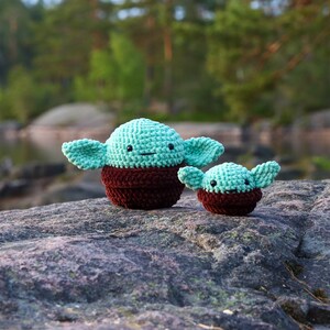 May include: Two crocheted plush toys shaped like a popular fictional character. The larger toy is green with brown accents and the smaller toy is green with brown accents. The toys are sitting on a grey rock.