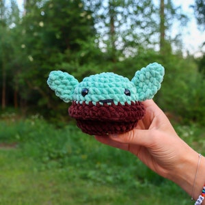 Pu&ograve; includere: Un giocattolo di peluche all'uncinetto a forma di un personaggio del franchise di Star Wars. Il giocattolo &egrave; verde con orecchie marroni e un viso marrone. Il giocattolo &egrave; tenuto in una mano.