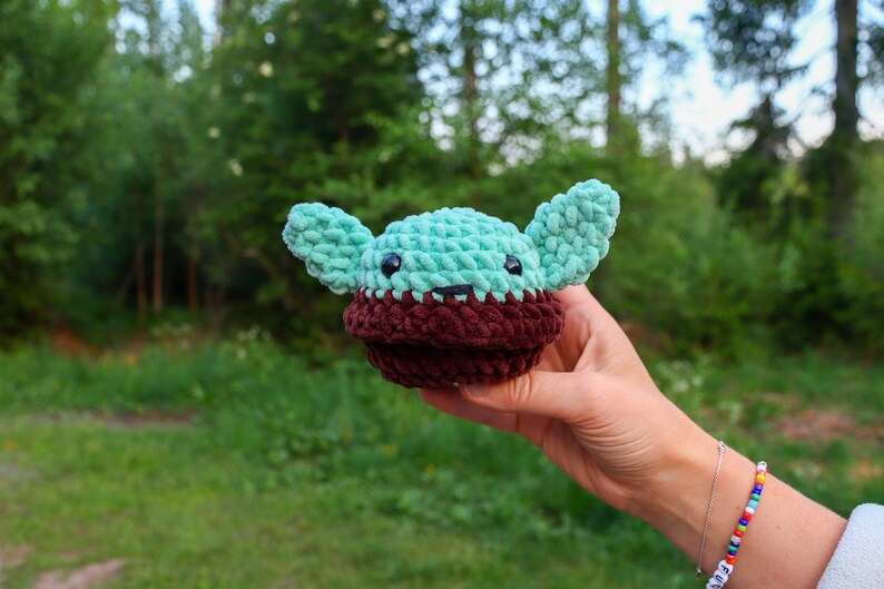 Pu&ograve; includere: Un giocattolo di peluche all'uncinetto a forma di un personaggio del franchise di Star Wars. Il giocattolo &egrave; verde con orecchie marroni e un viso marrone. Il giocattolo &egrave; tenuto in una mano.