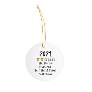 May include: White ceramic Christmas ornament with a gold string for hanging. The ornament has black text that reads "2021. Still Better Than 2020 but Still A Total Shit Show". There are four yellow stars above the text, representing a 4-star rating.
