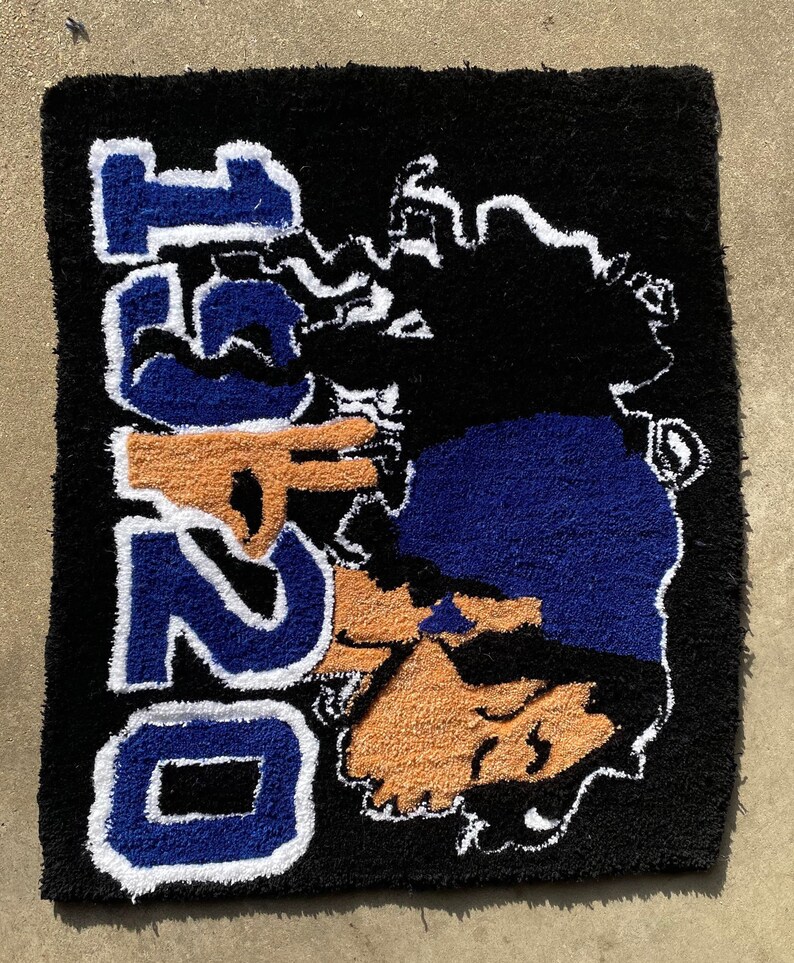 Puede incluir: Una alfombra negra con un dise&ntilde;o azul y blanco que dice "O251" y presenta una cara de dibujos animados con un sombrero azul y gafas de sol.