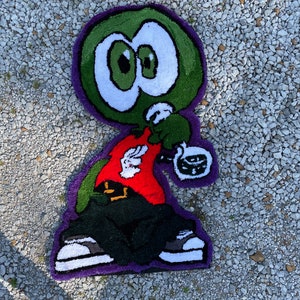 Puede incluir: Una alfombra de personaje de dibujos animados verde, rojo, negro y blanco con ojos grandes. El personaje lleva una camisa roja, pantalones negros y zapatillas blancas. La alfombra est&aacute; sobre una superficie de grava.