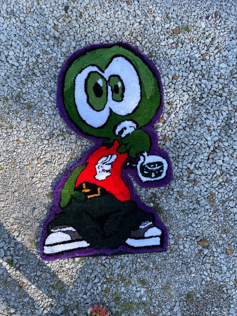 Puede incluir: Una alfombra de personaje de dibujos animados verde, rojo, negro y blanco con ojos grandes. El personaje lleva una camisa roja, pantalones negros y zapatillas blancas. La alfombra est&aacute; sobre una superficie de grava.