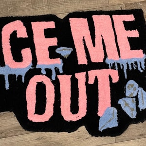Puede incluir: Una alfombra de bienvenida negra con las palabras "ICE ME OUT" en letras rosas. Las letras tienen una textura peluda y est&aacute;n delineadas en negro. Hay acentos azules y plateados alrededor de las letras.