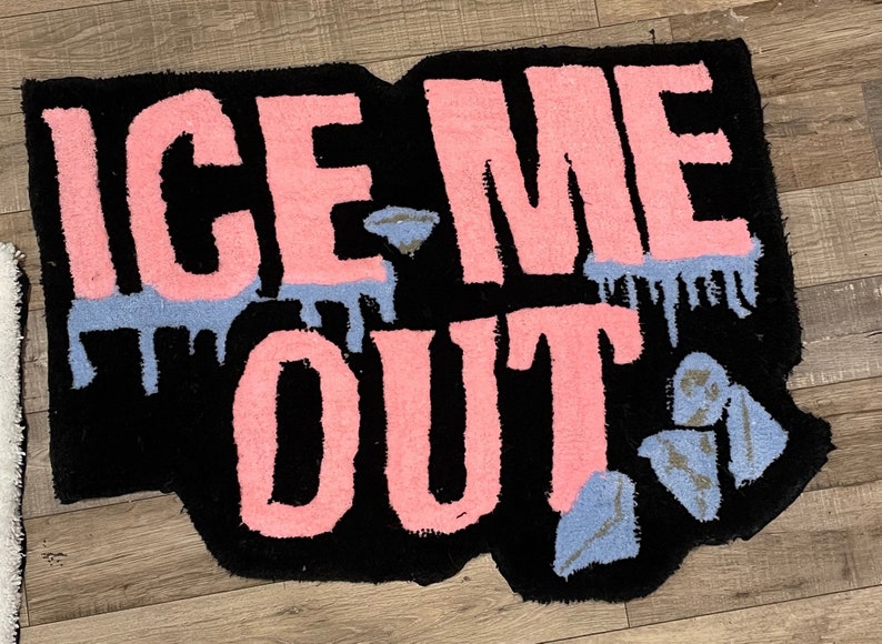 Puede incluir: Una alfombra de bienvenida negra con las palabras "ICE ME OUT" en letras rosas. Las letras tienen una textura peluda y est&aacute;n delineadas en negro. Hay acentos azules y plateados alrededor de las letras.