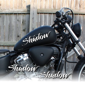 Könnte beinhalten: Schwarzes Motorrad mit verchromtem Auspuff und Lenker. Der Tank hat einen weißen "Shadow" Aufkleber. Das Wort "Shadow" ist auch in weiß auf dem Motor und unter dem Motorrad geschrieben.
