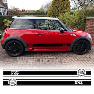 Può includere: Mini Cooper John Cooper Works rosso con strisce da corsa nere e una firma John Cooper Works. L'auto ha ruote nere e un logo Mini Cooper in bianco e nero.