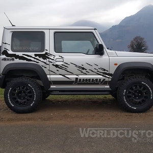 Może przedstawiać: Srebrny SUV z czarnymi oponami terenowymi i kołem zapasowym z tyłu. Pojazd ma czarne grafiki rozpryskowe i słowo "JIMNY" w kolorze czarnym. Tekst "4x4" jest widoczny z boku. Opony mają białe litery.