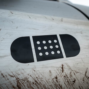 May include: A black and white band-aid sticker on a silver car door. The car door is covered in dirt and scratches.