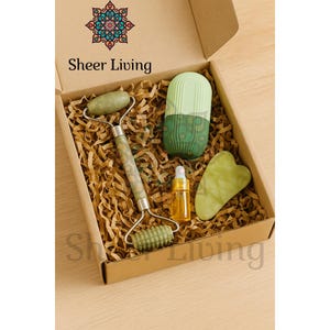 May include: A skincare gift set in a cardboard box, including a jade roller, a green and white ice globe, a small bottle of oil, and a green gua sha tool. The box is filled with brown paper and has the text "Sheer Living".