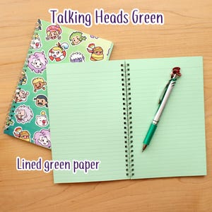 May include: A spiral-bound notebook with a green cover featuring cartoon characters. The notebook is open to reveal lined green paper. A green and silver pen with a red flower design rests on the open pages. The text "Talking Heads Green" and "Lined green paper" are visible.