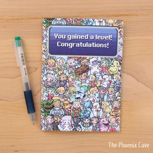May include: Greeting card with a colorful cartoon design and the text "You gained a level! Congratulations!". A clear pen with a green cap and blue grip rests on a wooden surface next to the card. The Phoenix Cave logo is at the bottom.