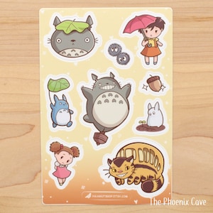 May include: A set of nine stickers featuring characters from the animated film My Neighbor Totoro. The stickers are on a white background with a yellow tint. The characters include Totoro, a cat bus, a girl with an umbrella, and a small white creature.