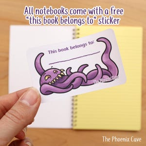 May include: A notebook with a free sticker. The sticker features a purple cartoon octopus with the text "This book belongs to:" and the shop name "The Phoenix Cave". The notebook is open to a blank white page.