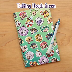 May include: A spiral-bound notebook with a green cover featuring cartoon character faces. The notebook is open, and a white and blue pen rests on the right side. The text "Talking Heads Green" is at the top.