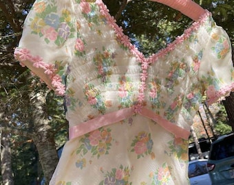 Puede incluir: Vestido vintage con estampado floral en colores pastel. El vestido tiene mangas cortas abullonadas y adornos rosas. Una cinta rosa define la cintura. El vestido está colgado de una percha rosa. El vestido mide unos 100 cm de largo.