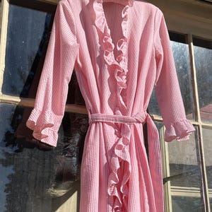 May include: A pink and white striped robe with ruffled trim and a matching belt. The robe has long sleeves with ruffled cuffs and is hanging on a hanger in front of a window.