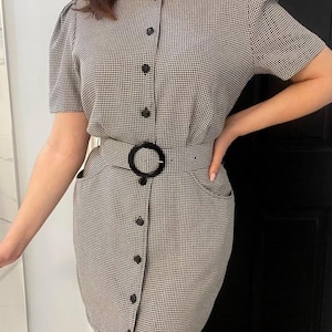Puede incluir: Un vestido a cuadros blanco y negro con mangas cortas y abotonado en la parte delantera. El vestido presenta un cinturón negro con hebilla circular y dos bolsillos delanteros. El vestido tiene cintura entallada y falda a la rodilla.