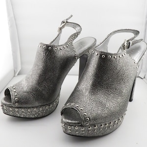 May include: A pair of silver platform heels with a peep toe and studded detailing. The heels have a slingback strap with a buckle closure.