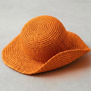 Peut inclure: Un chapeau de soleil orange vif doté d'une calotte arrondie et d'un bord large légèrement ondulé. Le chapeau est fabriqué à partir d'une spirale continue de matériau étroitement tissé, créant une surface uniforme et texturée sur l'ensemble.