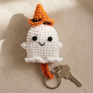 Peut inclure: Un porte-clés fantôme en crochet blanc avec un petit chapeau de sorcière orange à bande noire, deux yeux brodés noirs, un sourire cousu simple et un anneau métallique attaché en bas avec une clé argentée insérée dans l'anneau.