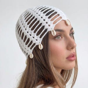 Peut inclure: Un bonnet en crochet blanc avec un motif ajouré et de petits coquillages cauris de couleur crème fixés le long du bord inférieur. Le couvre-chef est fabriqué en fil avec un motif géométrique répétitif créant des ouvertures verticales.