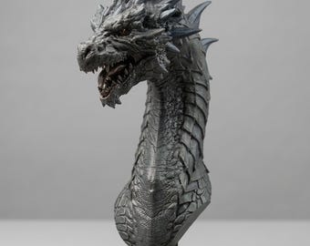 May include: A detailed dragon bust sculpture in shades of gray, with a textured, scaled appearance. The dragon's head is open-mouthed, revealing teeth. The sculpture sits on a dark wooden base, showcasing intricate details and a fantasy theme.
