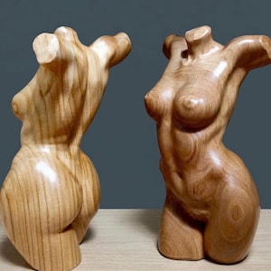May include: Two wooden sculptures of the female torso. The sculptures are carved from light-coloured wood, showcasing the natural grain. One sculpture is viewed from the back, the other from the front, both with smooth, polished surfaces. The sculptures are on a light-coloured surface.